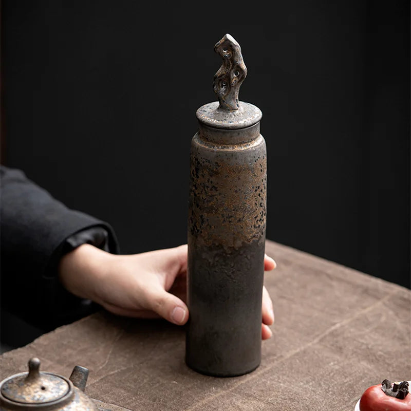 Hand holding a tall, dark grey ceramic storage jar with bronze accents on table