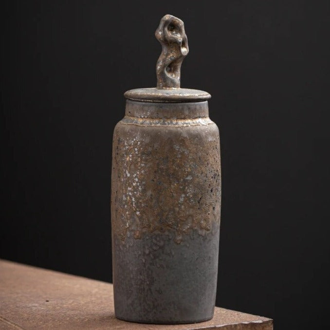 Dark grey ceramic storage jar with bronze accents and organic shaped handle on dark neutral background
