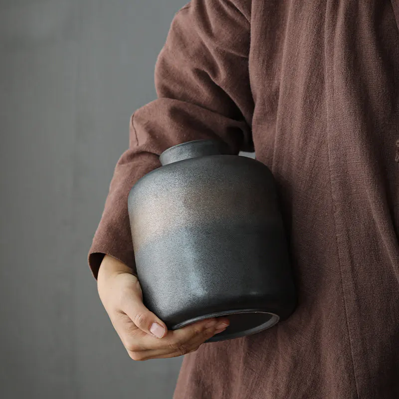 Hand holding dark grey ceramic storage jar with bronze accents on dark neutral background