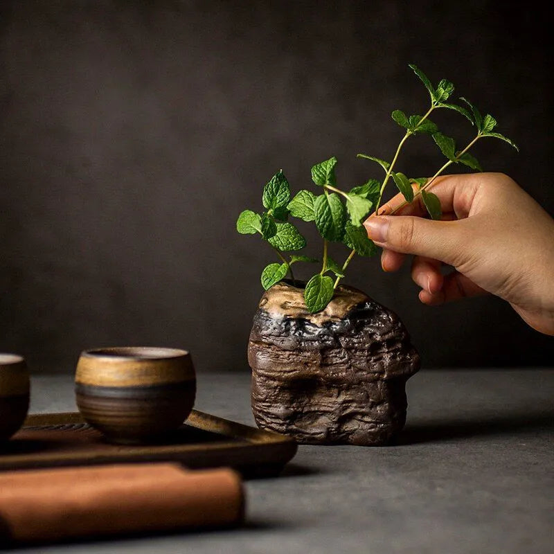 Hands putting herbs inside small, dark grey textured ceramic vase with bronze accents on dark neutral background