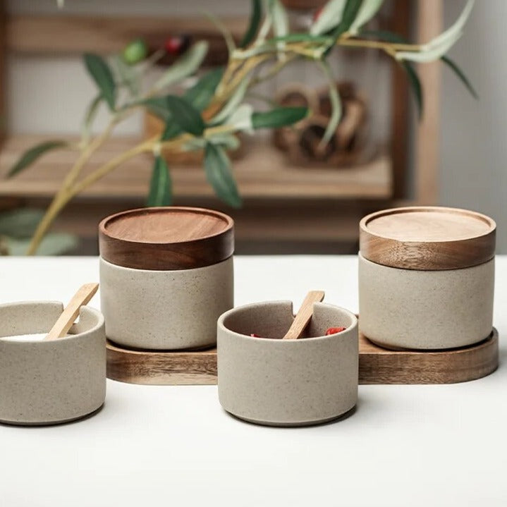 Set of grey spice jars with wooden lids, wooden spoons and oval wooden tray on white kitchen countertop