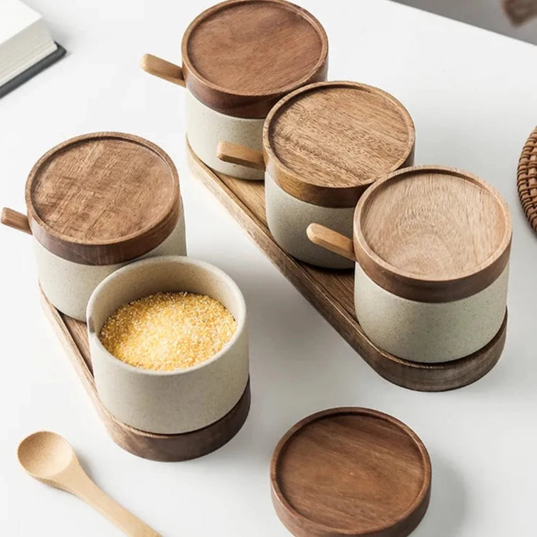 Elevated view of five grey spice jars with wooden lids, wooden spoons and oval wooden tray on white kitchen countertop