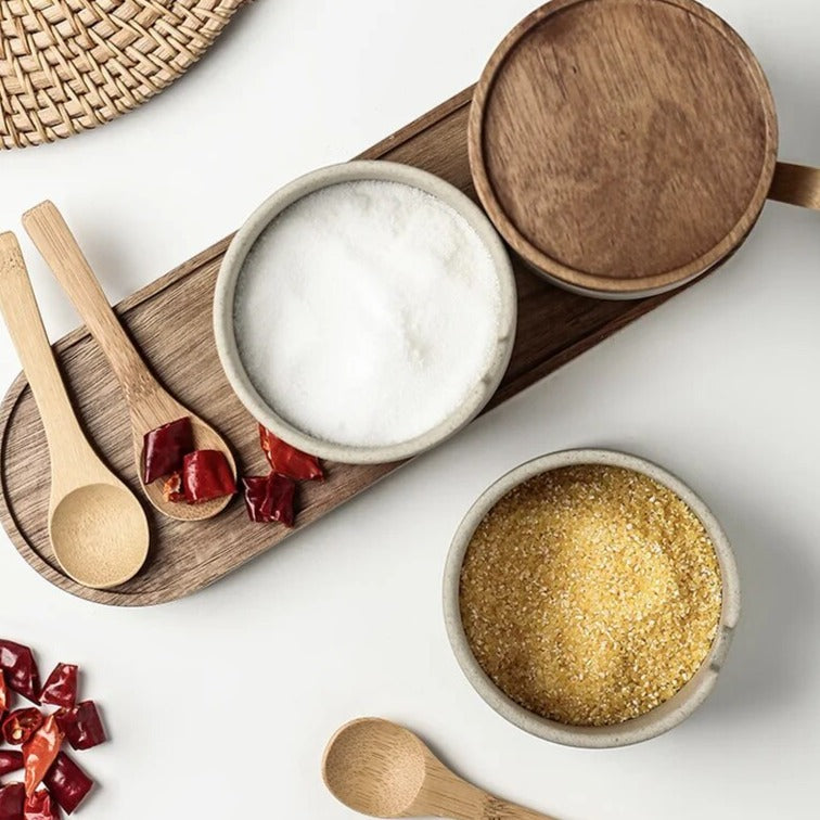 Top view of grey ceramic spice jars with wooden lids, wooden spoons and oval wooden tray on white kitchen countertop