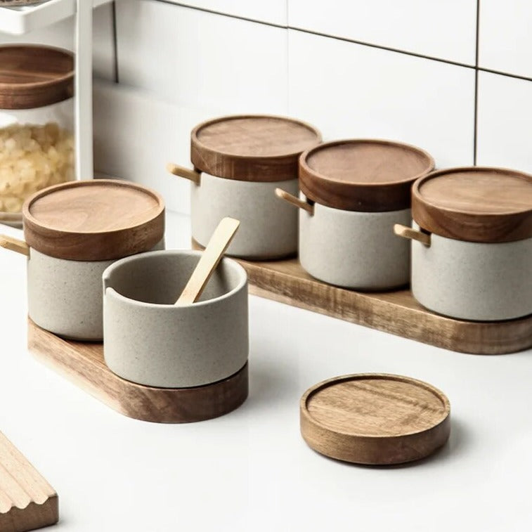 Set of grey ceramic spice jars with wooden lids, wooden spoons and oval wooden tray on bright kitchen countertop