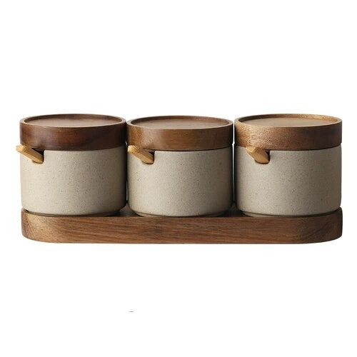 Set of three grey ceramic spice jars with wooden lids, wooden spoons and oval wooden tray on white background