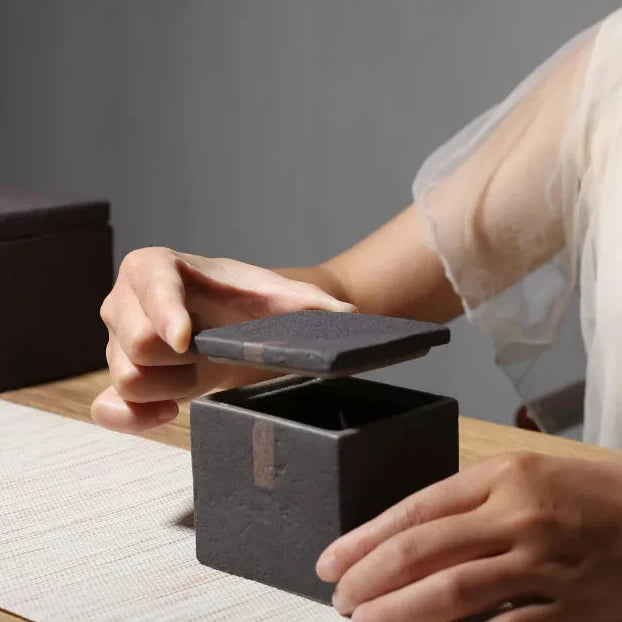 Woman's hands opening a dark grey textured square ceramic jar with decorative accent on wooden table