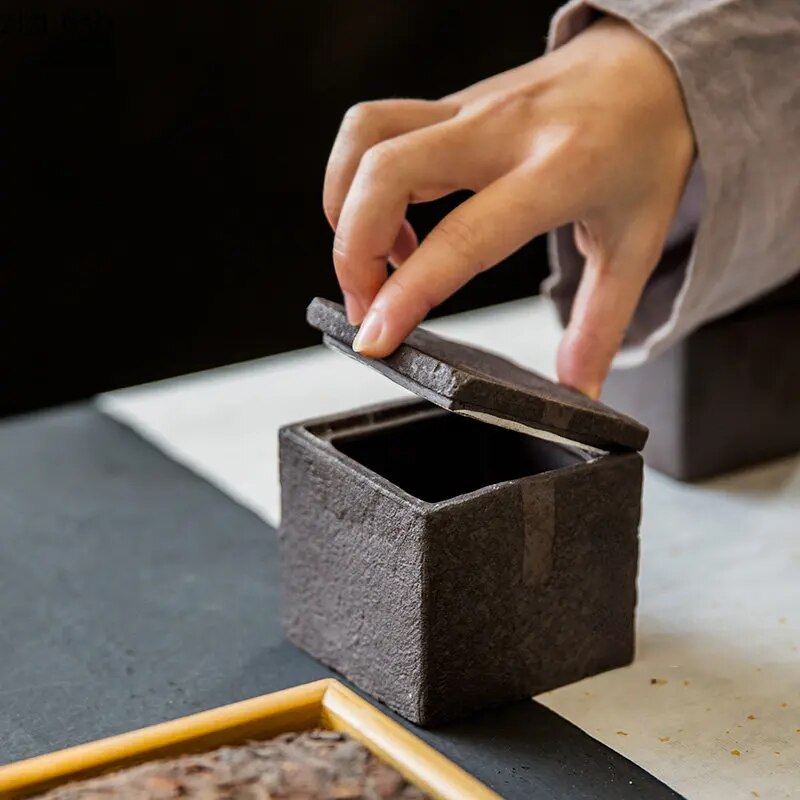 Woman's hands opening a dark grey textured square ceramic jar with decorative accent on table
